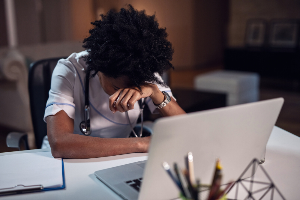 A veterinarian in scrubs is sitting at a desk with her head resting on her hand, appearing tired. In front of her is an open laptop, a stethoscope around her neck, and a clipboard on the desk. The background is dimly lit.